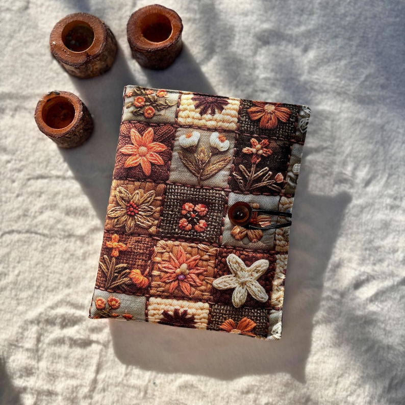 May include: A floral embroidered journal cover with a button closure. The cover features a patchwork design with various flower patterns in shades of brown, orange, and cream. Three small wooden candle holders are in the background.