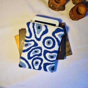 May include: A white pouch with a blue and light blue eye-like pattern. The pouch is made of a soft, textured fabric and has a zipper closure. The pouch is lying on top of two books.