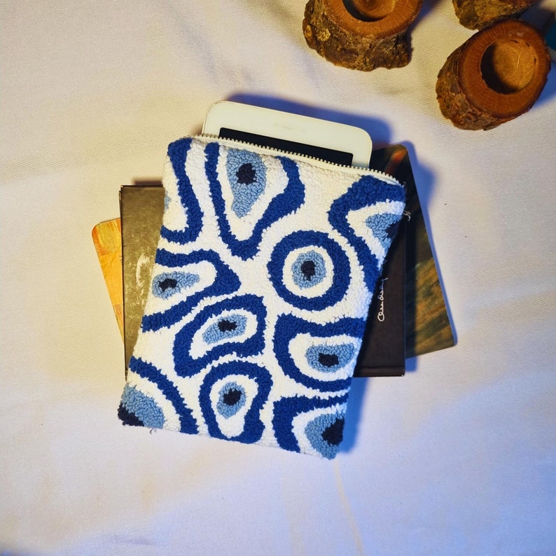 May include: A white pouch with a blue and light blue eye-like pattern. The pouch is made of a soft, textured fabric and has a zipper closure. The pouch is lying on top of two books.
