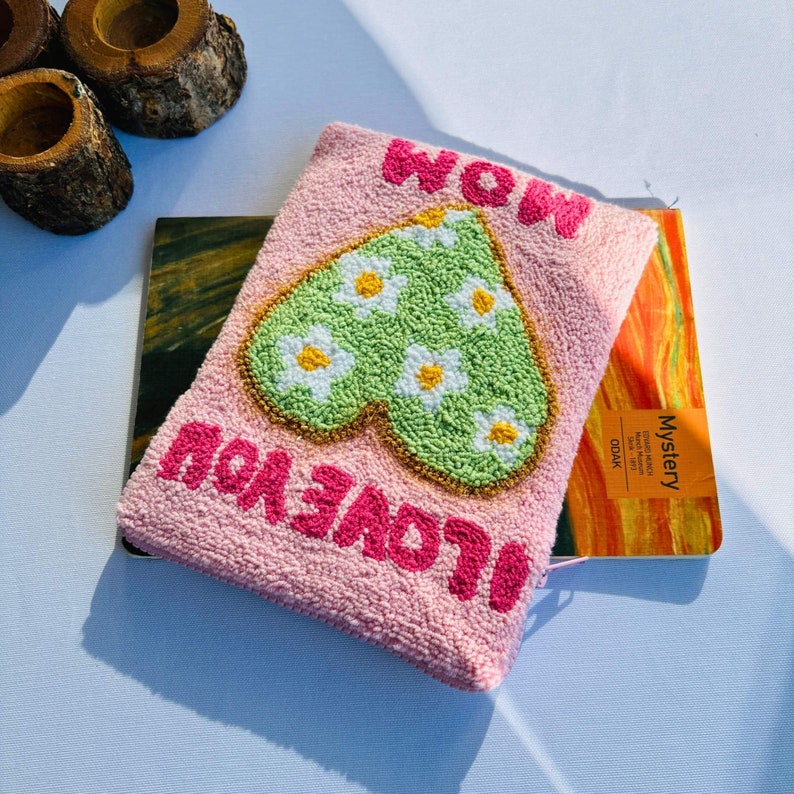 May include: A pink pouch with a green heart embroidered with white daisies. The pouch has the words "I LOVE YOU" and "WOW" written in pink.