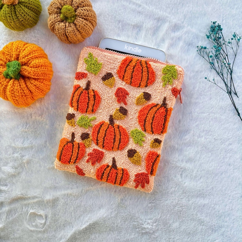 May include: A pink fabric pouch with a zipper closure, featuring a repeating pattern of orange pumpkins, green leaves, and brown acorns. The pouch is designed to hold a Kindle e-reader, and the word "Kindle" is visible on the e-reader.