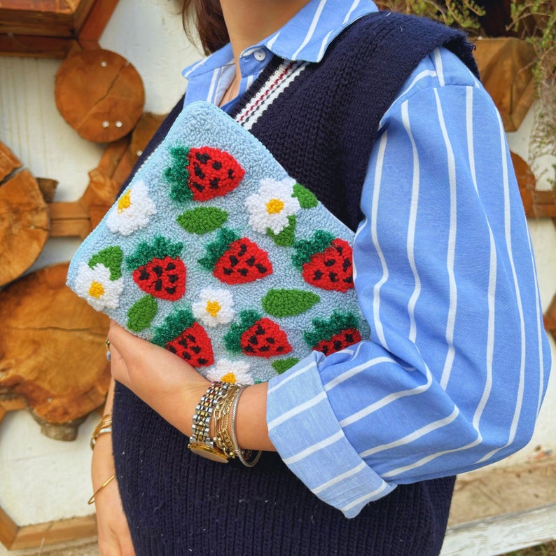 Peut inclure: Une chemise &agrave; rayures bleues et blanches avec un gilet en laine noir. La personne tient une pochette bleu clair avec un motif floral et des fraises.
