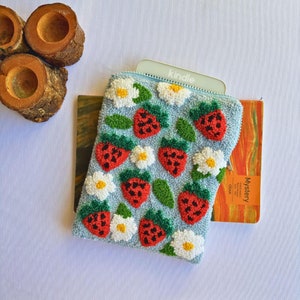 Peut inclure: Une pochette bleu clair avec une fermeture &eacute;clair orn&eacute;e d'un motif r&eacute;p&eacute;t&eacute; de fraises rouges et de fleurs blanches. La pochette recouvre partiellement une liseuse &eacute;lectronique grise avec le mot "kindle" imprim&eacute; dessus. La pochette est pos&eacute;e sur un livre dont la couverture porte le mot "Mystery".