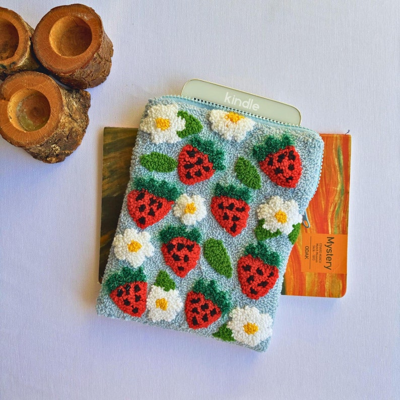 Peut inclure: Une pochette bleu clair avec une fermeture &eacute;clair orn&eacute;e d'un motif r&eacute;p&eacute;t&eacute; de fraises rouges et de fleurs blanches. La pochette recouvre partiellement une liseuse &eacute;lectronique grise avec le mot "kindle" imprim&eacute; dessus. La pochette est pos&eacute;e sur un livre dont la couverture porte le mot "Mystery".