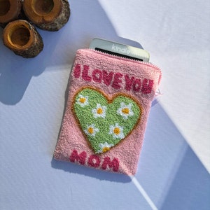 May include: Pink fabric pouch with a green heart with white flowers and the text "I LOVE YOU MOM" in pink. A silver Kindle e-reader is partially visible inside the pouch.