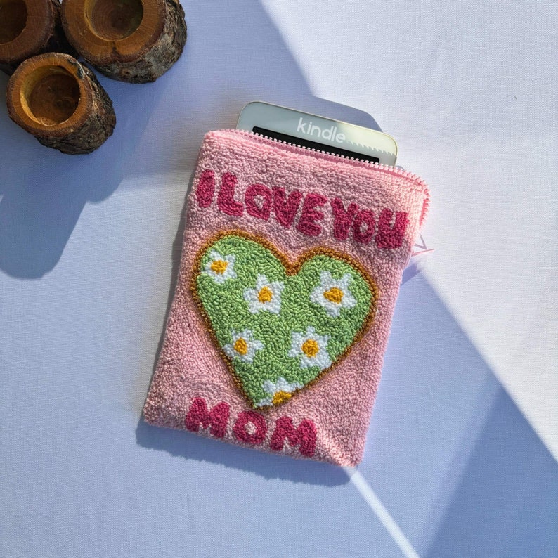 May include: Pink fabric pouch with a green heart with white flowers and the text "I LOVE YOU MOM" in pink. A silver Kindle e-reader is partially visible inside the pouch.