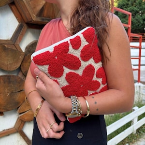 May include: A white clutch bag with a red floral design. The bag is made of a soft, textured fabric and has a zipper closure.