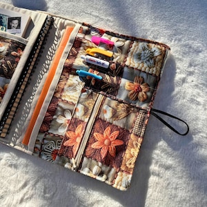 May include: A floral patterned journal cover with a variety of pockets and compartments. The cover features a patchwork design with brown, orange, and cream-colored floral patterns. Inside are notebooks, a pen, and a zipper pocket. The cover is closed with a black elastic band.