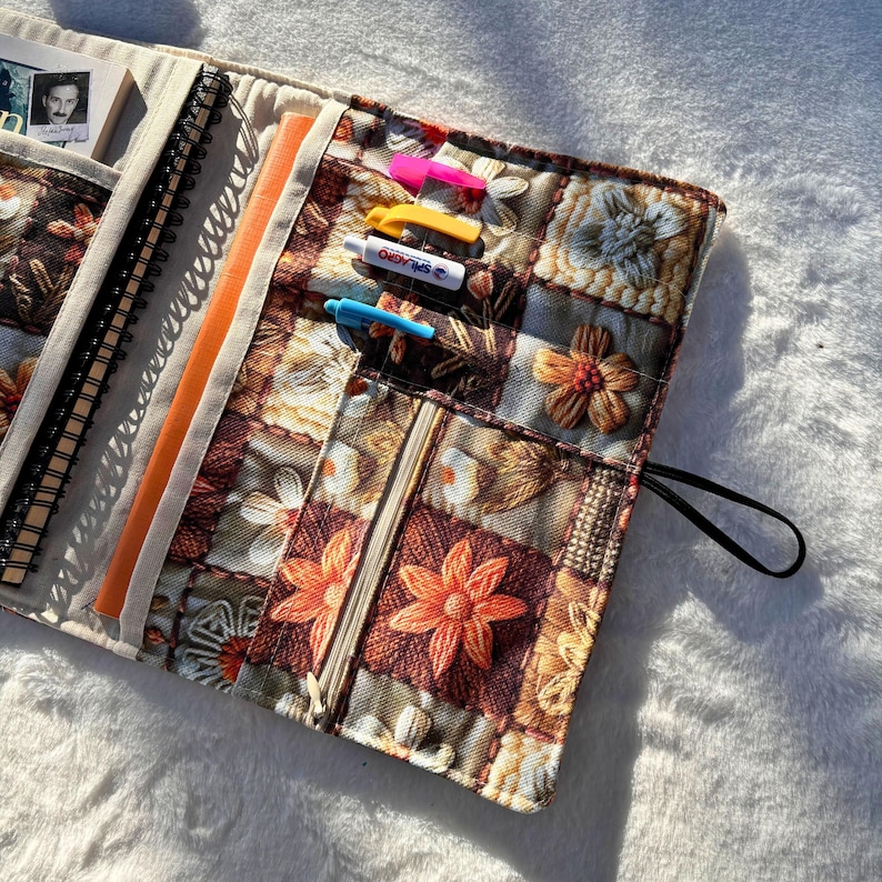 May include: A floral patterned journal cover with a variety of pockets and compartments. The cover features a patchwork design with brown, orange, and cream-colored floral patterns. Inside are notebooks, a pen, and a zipper pocket. The cover is closed with a black elastic band.