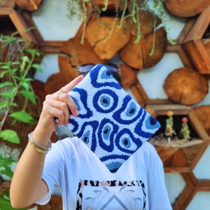 May include: A white and blue clutch bag with a swirling pattern of blue eyes. The bag is made of a soft, plush material and has a unique, eye-catching design.