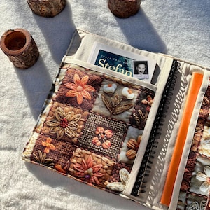 May include: A quilted journal cover with a floral patchwork design in shades of brown, orange, and cream. The cover has pockets holding a book and a notebook with an orange ruler. The journal is open, revealing the spiral binding.