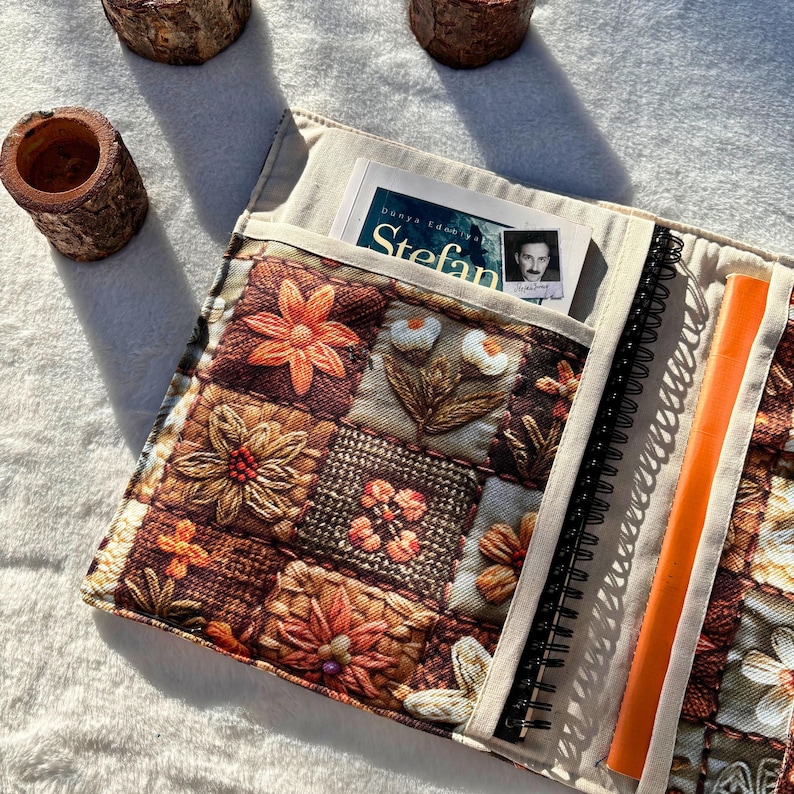 May include: A quilted journal cover with a floral patchwork design in shades of brown, orange, and cream. The cover has pockets holding a book and a notebook with an orange ruler. The journal is open, revealing the spiral binding.