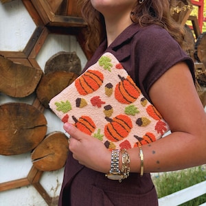 May include: A pink fabric clutch with an embroidered design of orange pumpkins, green leaves, and brown acorns.