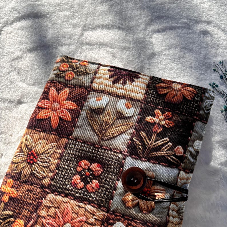 May include: A quilted book cover featuring a patchwork design with embroidered floral patterns in shades of orange, brown, and beige. The cover is secured with a dark brown button and a black elastic loop.