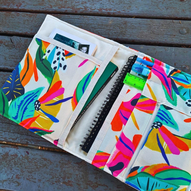 May include: A white canvas zippered pouch with a colorful floral print. The pouch has multiple pockets and is designed to hold a notebook, pens, and other supplies.