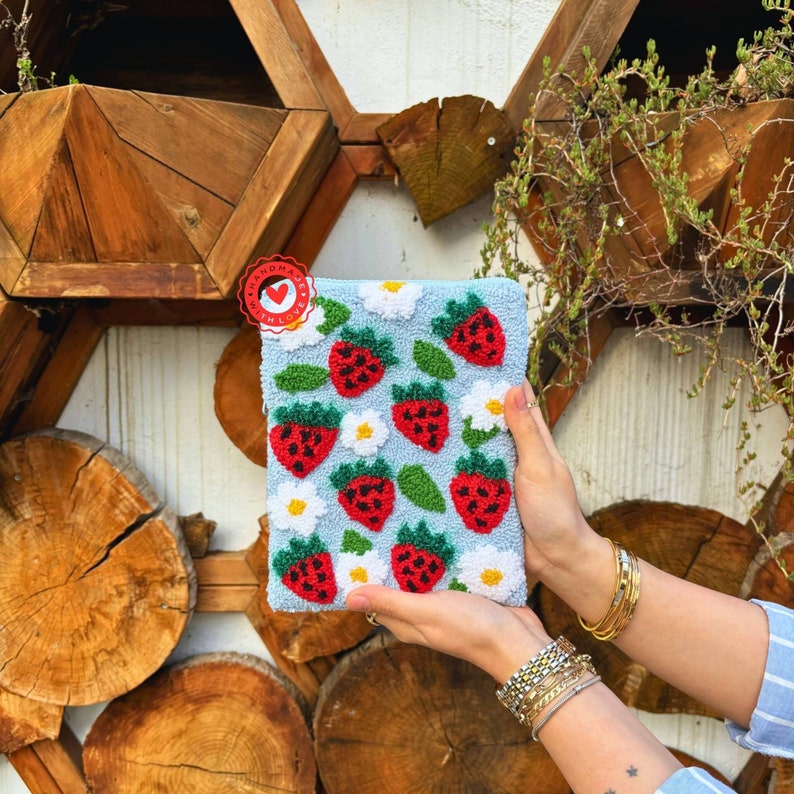 Peut inclure: Une pochette bleu clair avec un motif de fraises et de fleurs blanches cousu &agrave; la main. La pochette a un c&oelig;ur rouge avec le texte "Handmade with Love" dessus.