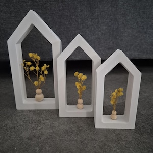 May include: Three white wooden house-shaped frames with arched tops, each containing a small wooden vase with dried yellow flowers.