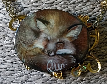 May include: A gold chain necklace with a round pendant featuring a painted image of a sleeping fox. The pendant is surrounded by decorative metalwork and has the word "Elen" inscribed on it.