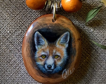 May include: A necklace with a wooden pendant featuring a hand-painted fox portrait. The pendant is attached to a black cord with brown wooden beads.