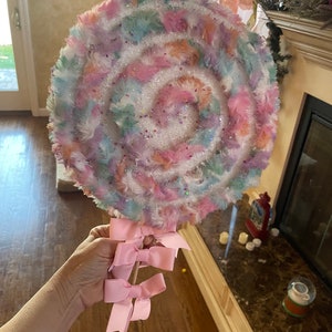 May include: A large, colorful, fuzzy lollipop with a white swirl design and pink bows attached to the stick.
