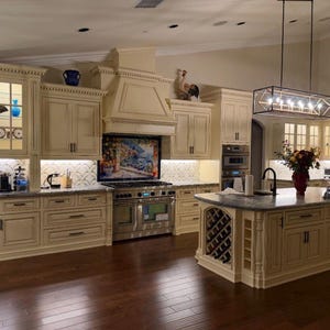May include: A kitchen featuring cream-colored cabinetry with dark hardware, a central island with built-in wine storage, a stainless steel range, a decorative backsplash mural, and a rectangular pendant light fixture hanging above the island.
