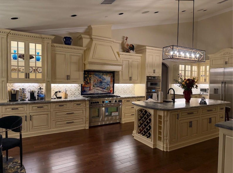 May include: A kitchen featuring cream-colored cabinetry with dark hardware, a central island with built-in wine storage, a stainless steel range, a decorative backsplash mural, and a rectangular pendant light fixture hanging above the island.