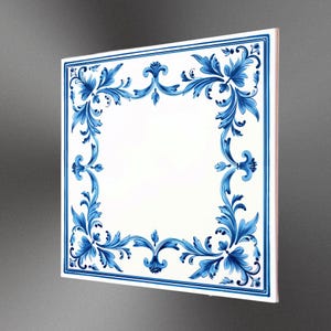 May include: Square white tile with a blue floral border. The design features ornate leaves and flourishes. The tile is angled, showing its thickness and a glimpse of the side.