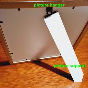 May include: A wooden picture frame with a white picture support and a black picture hanger. The frame is resting on a wooden surface. The picture support is angled, casting a shadow.