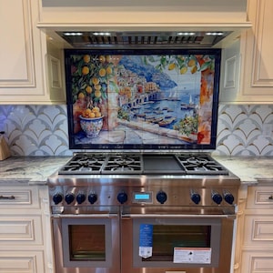 May include: A stainless steel dual-oven range with a central griddle and six burners, positioned beneath a decorative tile mural depicting a coastal Italian village with lemon trees, boats, and blue water, framed by cream-colored kitchen cabinetry.