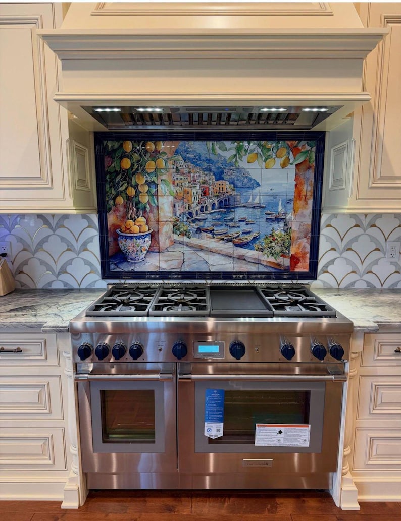 May include: A stainless steel dual-oven range with a central griddle and six burners, positioned beneath a decorative tile mural depicting a coastal Italian village with lemon trees, boats, and blue water, framed by cream-colored kitchen cabinetry.