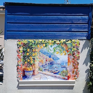 May include: A rectangular wall mural composed of ceramic tiles depicting a coastal Italian village scene with a lemon tree in a blue patterned pot, framed by a border of colorful mosaic tile shards, set against a blue horizontal wood panel.