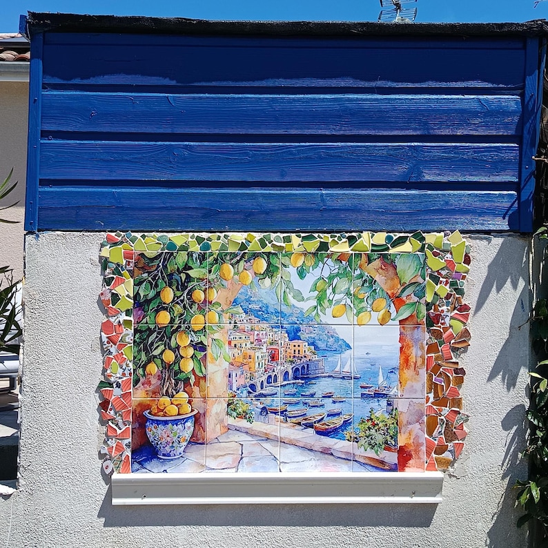 May include: A rectangular wall mural composed of ceramic tiles depicting a coastal Italian village scene with a lemon tree in a blue patterned pot, framed by a border of colorful mosaic tile shards, set against a blue horizontal wood panel.