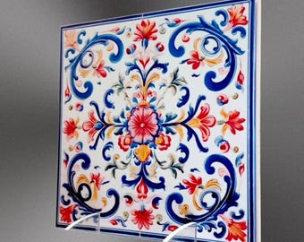 May include: A square ceramic tile with a white background and a colorful floral pattern. The pattern features blue, red, yellow, and green flowers and leaves. The tile is framed by a blue and white border.