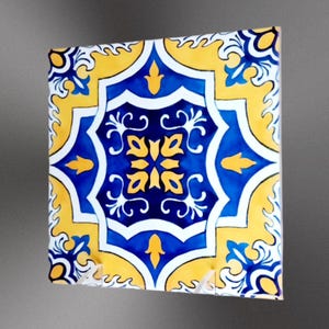 May include: A square ceramic tile with a blue and yellow geometric pattern. The tile has a white background with a blue center and yellow accents.
