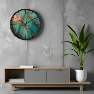 Puede incluir: Un reloj de pared redondo con marco negro. La esfera presenta un diseño abstracto en verde azulado y cobre. El reloj está montado en una pared gris sobre un mueble de madera con puertas grises. Una planta en maceta está a la derecha.