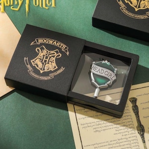 May include: A black box with a gold Hogwarts crest and the words "Hogwarts, Dormiens Nunquam Titillatus" on the lid. The box contains a silver pin with a green enamel shield that says "Head Girl".