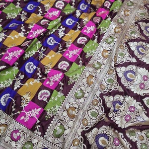 May include: A detailed shot of a maroon textile with a vibrant, geometric pattern. The design features squares in yellow, green, pink, and blue, each containing floral motifs. The border is adorned with intricate gold and silver embroidery, creating a luxurious look.