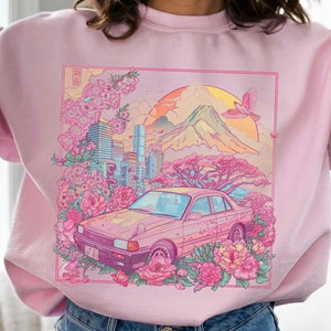 May include: A pink sweatshirt featuring a graphic print of a car driving through a city with cherry blossom trees and a mountain in the background. The graphic also includes a bird flying in the sky.