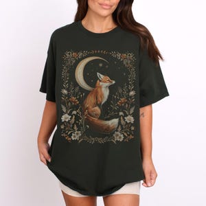 Op de afbeelding: Een donkergroen T-shirt met een grafische print van een vos die op een halve maan zit, omringd door bloemen. De vos is oranje en wit met een pluche staart.