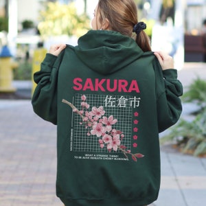 Puede incluir: Una sudadera con capucha verde oscuro con la palabra "SAKURA" en rosa en la parte superior de la espalda. Debajo, un diseño de cuadrícula presenta una rama de flor de cerezo y texto japonés. La frase "WHAT A STRANGE THING! TO BE ALIVE BENEATH CHERRY BLOSSOMS" está impresa en la parte inferior.