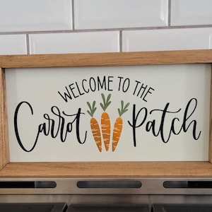 May include: A wooden sign with the text "Welcome to the Carrot Patch" in black lettering. Three orange carrots with green tops are depicted in the center of the sign.
