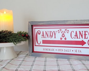 May include: A framed sign with a red background and white text that reads "CANDY CANES" with a red and white candy cane graphic. The sign also includes the text "HOMEMADE - OPEN DAILY - 50¢ EA."
