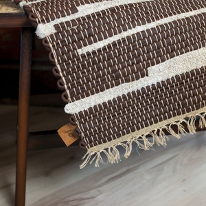 May include: A brown and white woven rug with fringe edges. The rug is made of a natural material and has a geometric pattern.