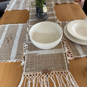 May include: A dining table setting featuring woven placemats and a matching table runner in shades of brown and white. A white bowl, plates, and a vase with greenery are on the table. The placemats have a fringed edge.