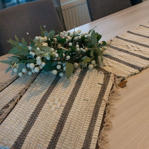 May include: Two white and gray woven table runners with fringe edges. The runners are on a light wood table with a white and green floral centerpiece.