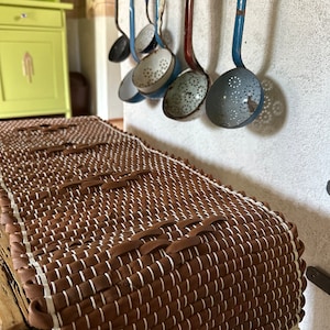 May include: A brown and white woven rug with fringe on the ends. The rug is made of leather and has a textured surface. The rug is hanging on a wall with a row of blue and red ladles.