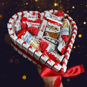 May include: A heart-shaped arrangement of assorted chocolate bars, including Kinder, Bueno, and Kinder Cards, decorated with red roses. The arrangement is held by a hand with a red ribbon.