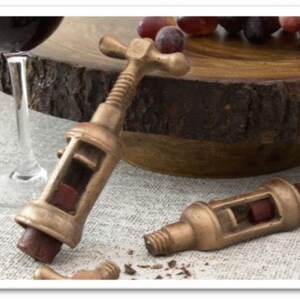 May include: Two antique-style bronze wine bottle openers with wooden handles. The openers are on a white linen surface with a wooden chopping board and a glass of red wine in the background.