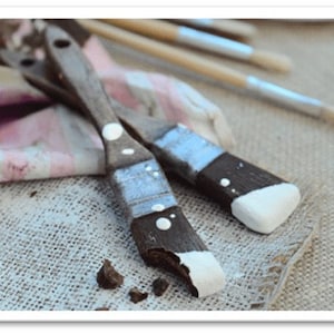 May include: Two paintbrushes with wooden handles and white paint on the bristles. The brushes are lying on a hessian surface.