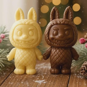 May include: Two whimsical figurines, one light yellow and the other chocolate brown, stand side-by-side. Each has a bunny-like hood and a smiling face. The figures are on a wooden surface, suggesting a festive or seasonal theme.